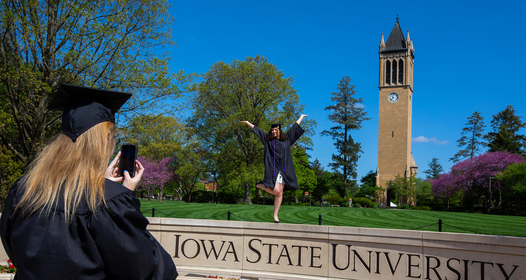 A graduate takes a picture of another graduate on the Iowa State University wall in front of the campanile